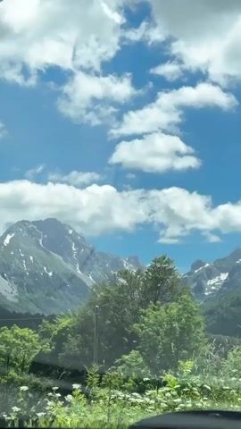 The video shows a scenic view from inside a vehicle. The foreground features the dashboard and part of the windshield, with a dark object, likely a dashcam, visible in the top left corner. Through the windshield, a mountainous landscape is depicted under a bright blue sky dotted with fluffy white clouds. Snow-capped peaks dominate the background, interspersed with green, tree-covered slopes in the middle ground. Some trees and foliage are visible closer to the vehicle, partially obscuring the view. Two small buildings with dark roofs are also visible nestled among the trees in the middle distance. The overall impression is of a sunny day in a mountainous region.