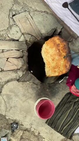 The video shows a traditional tandoor oven being used to bake flatbread. The oven is made of clay and stone, and it is filled with hot embers at the bottom. Several pieces of dough are attached to the inside walls of the oven, cooking and browning. One piece of bread is being turned with a metal skewer. In the foreground, a pink bucket and some fabric are visible. The overall scene suggests a rustic, outdoor cooking environment.