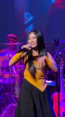 The video shows a live musical performance. A woman with long dark hair is singing into a microphone, wearing a yellow tube top with a black skirt and yellow arm warmers. She is on stage with a band. In the background, a person wearing a red hat is playing drums, and another person with long dark hair is playing an instrument. The stage is lit with purple and blue lights. The woman is gesturing with her left hand as she sings.
