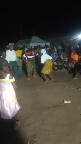 A group of people are gathered outdoors at night, around a dirt area. Several individuals are dancing in the center of the area, including three women wearing patterned skirts and tops, and one woman in a white top and green skirt. A young girl in a pink dress stands to the left, facing away from the camera. A crowd of onlookers is visible in the background, some sitting and others standing. There are lights illuminating the area, and a tent is set up behind the dancers.