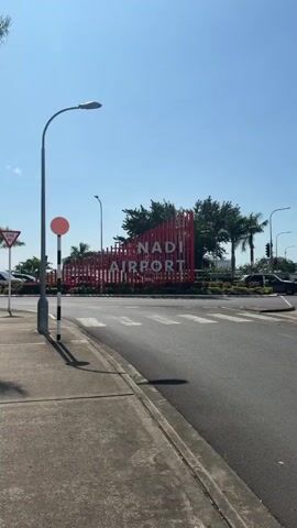 The video captures the exterior of Nadi Airport in Fiji on a clear, sunny day. The camera pans from left to right, starting with a sidewalk and a street. A prominent red sign with white lettering reads "NADI AIRPORT." Palm trees and other tropical foliage are visible in the background. Several flags are flying, including the Fijian flag and what appears to be an airline or airport service flag. The sky is blue with some scattered clouds. The overall environment suggests a warm, tropical climate typical of an airport setting. No people are clearly visible in the video.