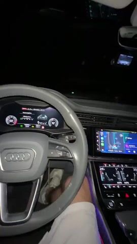 A person is driving an Audi car at night. The car's interior lights are on, illuminating the dashboard and the driver's hands on the steering wheel. The car's speedometer shows 102 km/h. Another person is sitting in the passenger seat, talking and looking towards the driver. The video cuts to a view of the road ahead, then back to the car's interior, showing the driver adjusting the settings on the dashboard.