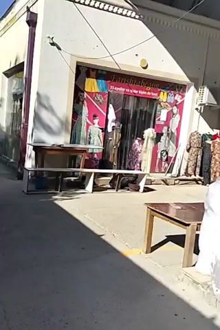 The video shows an outdoor scene, likely a marketplace or a street with shops. In the foreground, there is a wooden table with a white top, and another long, low white shelf unit with items underneath. A white plastic bag is visible on the right side of the frame.

The main focus is a storefront with a large glass window displaying clothing on mannequins and racks. The shop's name, "Farishotegym," is visible above the window, along with some text in what appears to be a foreign language. The window display includes several dresses on mannequins, some of which are floral patterned. There are also images of models wearing dresses on the exterior of the storefront. To the right of this shop, another storefront is partially visible, with some greenery and text above its entrance.

The ground is paved with a concrete-like material, and shadows from the structures indicate sunlight. The overall impression is of a commercial area on a sunny day. There are no people clearly visible interacting within the frame, though there might be figures in the shop windows. The video appears to be a static shot or a very slow pan.