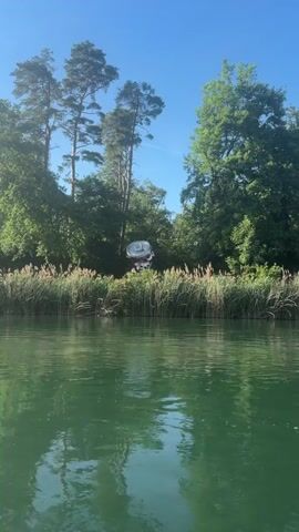 A drone flies over a body of water and towards a treeline with reeds in the foreground. A large, crumpled aluminum can is suspended in the air above the reeds, attached to a multicolored, possibly fabric, structure. The drone is not visible in the frame. The sky is clear and blue.