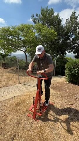 A man wearing a white baseball cap with "COLLECTIVE MEMORY" and "CODE: T8X33" printed on it, is exercising outdoors on a red piece of fitness equipment. The equipment appears to be a stair stepper or similar device. He is dressed in a grey polo shirt and dark pants, with a smartwatch on his left wrist. The man is actively engaged with the machine, gripping the handlebars and pushing down with his legs.

The setting is outdoors, with dry, straw-like ground cover around the fitness equipment. Behind the man, there is a metal fence and a path. Lush green trees are visible in the background, and beyond them, a scenic view of rolling hills and a distant landscape. The sky is blue with scattered white clouds, suggesting a clear and sunny day. The time of day appears to be midday or early afternoon. The man's focus and exertion suggest he is engaged in a physical workout. The location is identified as Misgav Regional Council, Israel.