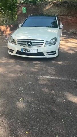 A white Mercedes-Benz C-Class sedan is parked in an outdoor paved area. The car is viewed from the front, with the license plate KP61 FCC clearly visible. The vehicle has tinted windows and a black roof. Behind the car, there is a brick wall and some greenery. The ground in front of the car appears to be asphalt or tarmac. Sunlight casts shadows on the ground.