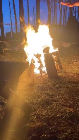 A bonfire burns brightly in a grassy area at dusk. The flames are tall and active, illuminating the surrounding dry grass and dirt. Several logs are stacked in the center of the fire. To the left, a dark, metallic object, possibly a chair or part of a grill, is partially visible in the foreground, casting a warm glow. In the background, tall, thin trees are silhouetted against a darkening blue sky. A person’s hand is briefly visible reaching towards the fire from the right side of the frame, and a pair of feet in sandals are also visible near the fire. Some indistinct structures or objects are visible in the far background, suggesting an outdoor gathering or campsite. The overall atmosphere is warm and inviting.