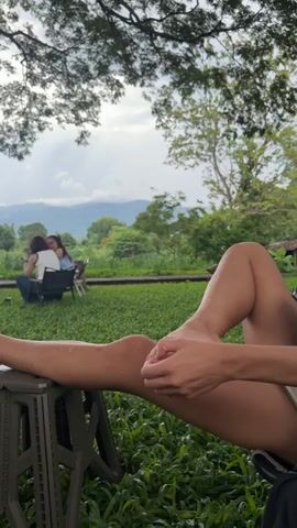 The video shows a person's leg propped up on a stool in the foreground. The person's leg is slightly bent at the knee and their hand is resting on their shin. In the background, two people are sitting at a table in what appears to be a park or garden. The environment is lush and green with trees and hills in the distance. The sky is cloudy.