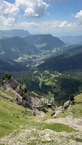 This is a vertical, first-person perspective video from a moving cable car ascending a mountain. The cable car is not visible in the frame. In the foreground, a steep, grassy mountainside with rocky outcrops dominates the left side of the screen, while the right side shows more rocks and sparse vegetation. Multiple parallel black cables stretch diagonally across the upper left portion of the frame, disappearing out of view. In the middle ground, a cable car cabin, dark gray and rectangular, hangs suspended from the cables, descending into the valley. Below the cable car, a lush green valley unfolds, dotted with scattered trees and patches of grass. A winding road can be seen traversing the valley floor. Further in the distance, a small town or village is nestled within the valley. The background is filled with layers of majestic, hazy blue mountains under a bright, partly cloudy sky. The overall impression is one of a scenic mountain ascent, offering a sweeping panorama of the valley and surrounding peaks.