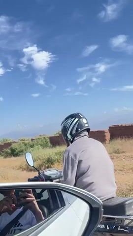 The video shows the view from inside a car driving on a road. In front of the car, a man is riding a small motorcycle. The rider is wearing a gray sweater, striped pants, a black helmet with white and gray stripes, and sandals. He has a bag strapped to the back of his motorcycle. On the side of the road, there is dry brush, remnants of brick structures, and trees. In the distance, mountains can be seen under a bright blue sky with scattered clouds. The car's side mirror is visible in the bottom left of the frame, reflecting the person recording the video who is wearing a white shirt and holding a phone. The motorcycle has a license plate that reads "26 43794".