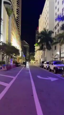 The video depicts a street scene at night, possibly in a city like Miami, based on the presence of palm trees and architecture. The street has parked cars on one side, including Jeeps and a Tesla. People are walking on the sidewalks and across the street, including a person pushing a stroller. A brightly lit Publix supermarket can be seen in the background. The lighting suggests a mix of streetlights and illuminated building facades. A white arrow on the street indicates the direction of traffic. The overall atmosphere suggests an urban environment with typical nighttime activity.