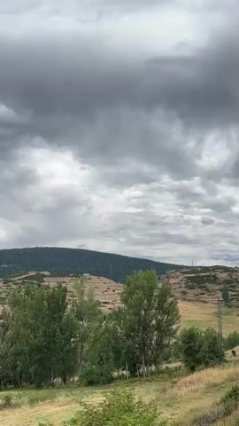 A landscape video shows a cloudy sky over rolling hills and trees. The clouds are dark and appear to be moving. The hills are covered in dry grass and some trees. In the foreground, there are several trees with green leaves. The overall impression is of a somewhat overcast or potentially stormy day in a rural area.