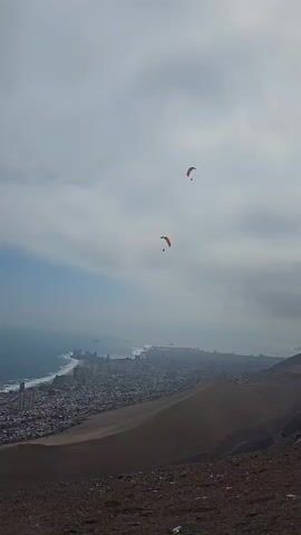 Two paragliders are flying against a grey, overcast sky. The paragliders are red. Below them is a city on a coastline.  A large sand dune is in the foreground. The camera zooms in on one of the paragliders.  The sky is mostly grey.