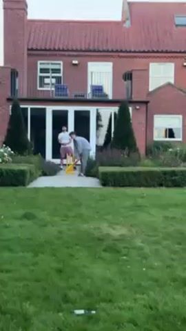 The video shows a group of young men playing cricket in a backyard. The batsman is wearing a light-colored shirt and gray pants. He swings the bat, hitting a yellow ball. Several other men are watching the play. One of them is wearing a white shirt, another one is wearing a light blue and white soccer jersey with the number 10 on it. The backyard has a well-maintained lawn, hedges, and several small trees. A large red brick house with white framed windows and doors is in the background. There is a bottle lying on the lawn in the foreground.