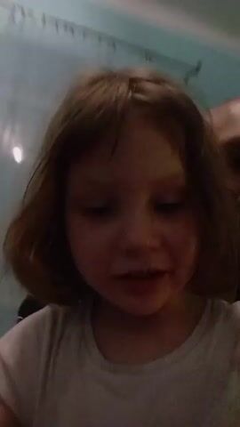 A young girl with short brown hair is in the foreground, with a bald man behind her, slightly out of focus. The girl is wearing a plain white t-shirt and smiles at the camera. She waves her hand, then crosses her arms. The man, wearing a black and white striped shirt, smiles at the camera. A light turquoise wall and partially seen white curtain are in the background.