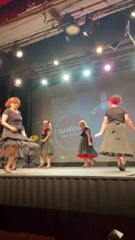 A group of women in black dresses and red accents are dancing on a stage. The stage has a large screen behind them displaying a logo that reads "Aula de Baile" and "Songs of." Lights are focused on the performers, and some lighting equipment is visible above the stage. The women are wearing similar outfits, including full skirts that swirl as they dance. One woman on the far left has a red ribbon in her hair. Another woman in the center has a red sash around her waist. The women are all facing different directions as they perform synchronized movements. Some of the women have their arms outstretched. There is a drum set visible on the left side of the stage. The overall impression is a performance or rehearsal.