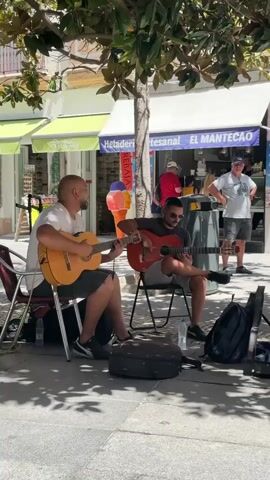 A sunny day outdoors with two men playing acoustic guitars on a sidewalk in front of shops. The man on the left is wearing a white t-shirt and dark shorts and is seated on a red chair. The man on the right is wearing a dark t-shirt and shorts and is seated on a folding chair. Their guitars are visible. There are bags on the ground near them, possibly for donations. Behind them, there is an ice cream shop with colorful signage. A few other people are in the background, including a man standing near the ice cream shop entrance. A woman is partially visible in the foreground on the right side of the frame, holding a white bag. The ground is paved, and there are trees providing shade.