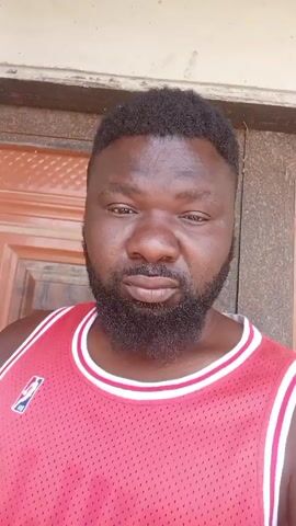 The video shows a man wearing a red and white basketball jersey standing in front of a brown door. The man is dark-skinned and has a full beard and short, curly hair. He is making various gestures with his hand, including forming a peace sign, rubbing his beard, and placing his hand on his throat. The background consists of a wooden door with panels and what appears to be a white or light-colored wall. The lighting appears to be natural daylight.