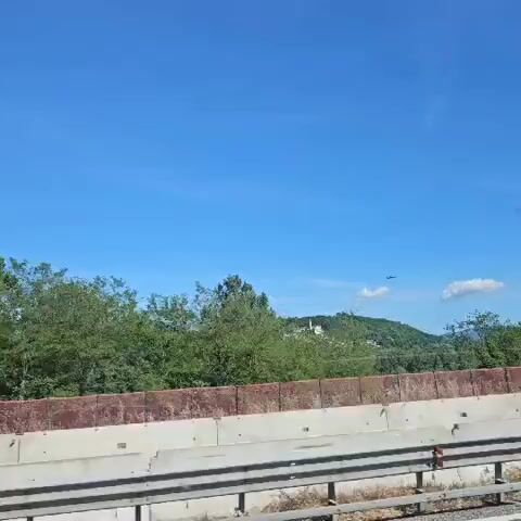 The video shows a highway driving scene on a clear, sunny day. A helicopter flies in the distance over a small town on a hill, featuring a large building that could be a castle or fortress. The camera passes by trees, a barrier, and a concrete edge on the highway. The overall impression is of a typical road trip with some scenic views.