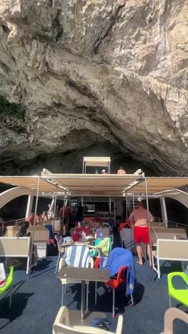 A crowded tour boat navigates through a narrow sea cave. Sunlight filters through the opening of the cave, illuminating the boat and the rough, textured rock walls. People are seated on benches and chairs on the open deck of the boat, some appear to be wearing swimwear and have towels draped over their laps or on chairs. A man in red swim trunks walks across the deck. Various objects, including bags, towels, and what appear to be cups and bottles, are scattered on tables and chairs. The boat's canopy provides shade for some areas of the deck. The overall impression is a lively, tourist excursion within a natural geological formation.
