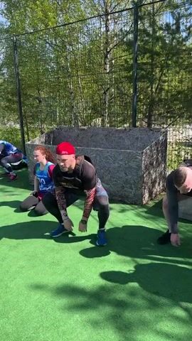 Here is a description of the video:

The video shows four people doing squats on an artificial turf field. Three men are visible, and one woman is kneeling in the background. The men are wearing weighted vests and shorts or leggings, and they appear to be exercising together. The field is surrounded by a chain-link fence and trees. There are water bottles and other personal items on the ground near where they are exercising. The sun is shining brightly, casting shadows on the field.