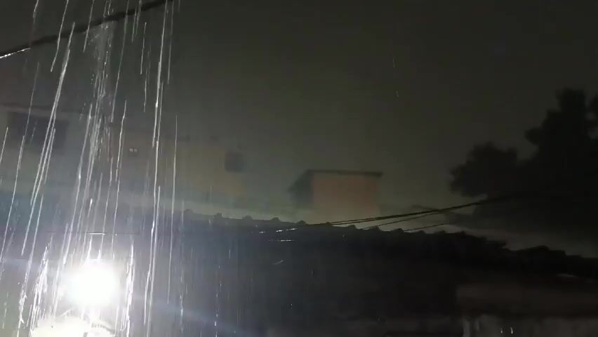 Here is a concise description of the video:

The video depicts a heavy downpour at night. Rain streams down intensely, illuminated by a bright light source in the foreground. In the background, buildings are visible, though shrouded in darkness and potentially fog. A section of a rooftop is also in view, and the general atmosphere appears to be gloomy and wet.