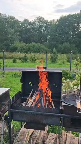 A campfire burns brightly in a metal barbecue grill. The fire consists of several pieces of wood, with bright orange flames licking upwards. The grill is set on a black metal stand. In the background, there is a green lawn, some trees, and a cloudy sky.
