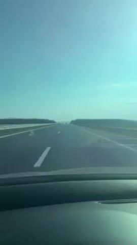 The video shows a car driving down a straight highway under a clear, light blue sky. The road is asphalt, with white lane markings. Trees line either side of the highway. No other vehicles are visible nearby. The video is taken from inside the car, showing the dashboard. The horizon is visible in the distance.
