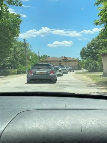 The video shows a row of cars driving through a small village. The view is from inside a car, looking out through the windshield. The car in front of the viewer is a dark gray Fiat Abarth with Bulgarian license plates. The other cars in the line are mostly silver or light-colored. There is a house with a red tile roof and a few trees to the side of the road. The sky is blue with some fluffy white clouds. A Bulgarian flag hangs from a utility pole on the left side of the road. A techno song is playing in the background.