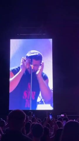 A large screen displays a man wearing a black t-shirt with red lettering that reads "SIN" in a concert setting. The man is holding a microphone and singing. The screen is illuminated by stage lighting, which casts red and blue hues. Below the screen, a crowd of people can be seen, many holding up their phones to record the performance. The overall atmosphere appears to be that of a live music event.