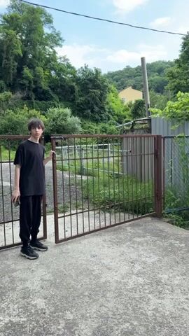 A young person stands to the left of an open, rusty metal gate. They are wearing a black t-shirt, black pants, and sneakers, and holding a dark object, likely a phone, in their left hand. The gate is made of vertical metal bars and appears to be a double gate, with one side slightly ajar. The ground in front of the gate is a concrete surface.

Behind the gate, there is an area with overgrown green grass and some vegetation. Further in the background, lush green trees cover a hillside. A yellow house with a reddish roof is visible on the hillside through the trees. A tall, grey cylindrical object, possibly a utility pole, stands on the hill. To the right of the gate, a corrugated metal fence runs parallel to it. The overall scene suggests a somewhat neglected outdoor area.

The lighting indicates it is daytime, with the sun casting a slight shadow. The sky is blue with some scattered clouds, suggesting pleasant weather. The presence of greenery and the overall outdoor setting point to a possibly rural or suburban environment. The young person's expression is neutral, and their posture is relaxed as they stand beside the gate.

No visible text is present in the media file.

The location context is Shepsi, Russia.
