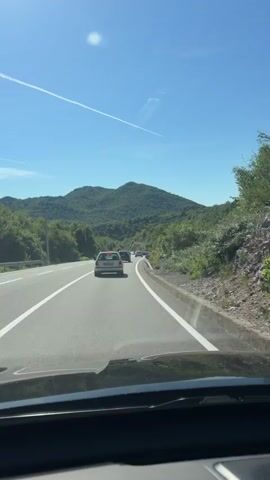 The video captures a first-person perspective of driving on a highway on a clear, sunny day. The road is paved and has multiple lanes, indicated by white lines. Other cars are visible on the road, driving in the same direction. The highway is bordered by a metal guardrail on the right and lush green trees and vegetation on both sides. In the background, rolling hills covered in dense green foliage rise against a bright blue sky, which features a faint contrail from an airplane. The dashboard of the vehicle is partially visible at the bottom of the frame.