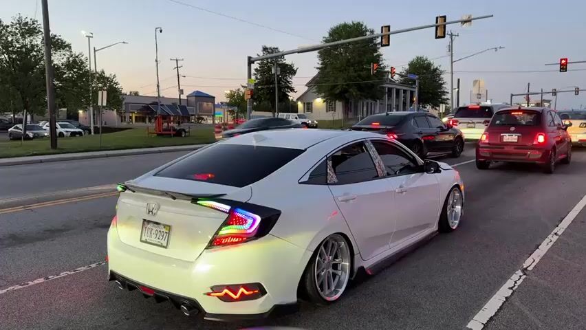A white Honda Civic is stopped at a traffic light on a busy street. The car is heavily modified with custom taillights and a spoiler. There are other cars waiting at the light, including a black sedan and a red Fiat. Cars pass in the opposite direction. The sky is a dusky blue, indicating it is either early morning or late evening. There are buildings and trees in the background. The license plate of the Honda Civic is visible and reads "TUX-9297." The sound of an engine revving can be heard.