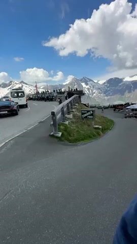 A first-person perspective video shows a couple taking a selfie in the mountains. The man and woman are wearing sunglasses. Behind them, a wooden building with the name "EDELWEISS HUTTE" is visible. The landscape is mountainous with snow-capped peaks and a blue sky with clouds. A road curves around the building.