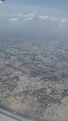 The video shows an aerial view of a landscape from an airplane window. The ground below is a patchwork of agricultural fields, some appearing fallow and others with crops. Scattered throughout the fields are clusters of buildings, suggesting villages or small settlements. The overall impression is of a rural area viewed from a high altitude. The sky above is partially obscured by clouds, and the light suggests it is daytime. The edge of the airplane's wing is visible in the lower left corner of the frame.