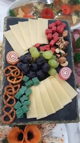 This is a close-up, high-angle shot of a cheese platter. The platter is a rectangular black slate, adorned with a variety of food items. The main subjects are the food items themselves: thinly sliced light yellow cheese, arranged in a fan shape; small, cubed red candies; a mix of nuts including walnuts and cashews; dark purple and green grapes; miniature pretzels; and small, cube-shaped green candies. There are also two swirl-shaped candies, one white with a red swirl and the other pink with a white swirl, placed on either side of the platter.

The platter is likely a snack or appetizer, possibly for a party or gathering. The arrangement of food suggests it is a prepared dish for consumption. The lighting is bright and even, indicating it was likely taken indoors, possibly in a restaurant or at a catered event. There are no people visible in the frame. The background is blurred but suggests more food items, possibly also part of a larger spread, with hints of red and white decorative elements that might be garnishes or part of table settings. The overall impression is one of a well-presented and appetizing food display. No text is visible in the image.