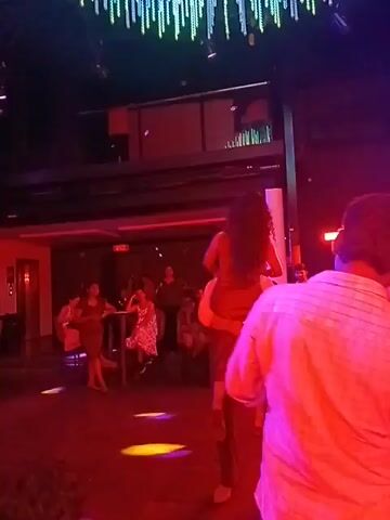 The video shows a crowded nightclub with colored lights and a decorative ceiling fixture made of dangling strands.  A man is holding a woman in his arms, raising her up so she can spread her arms wide. Other people stand around the dance floor, some watching the couple and some sitting at tables.  The nightclub appears to be dimly lit, with red and blue lighting effects.