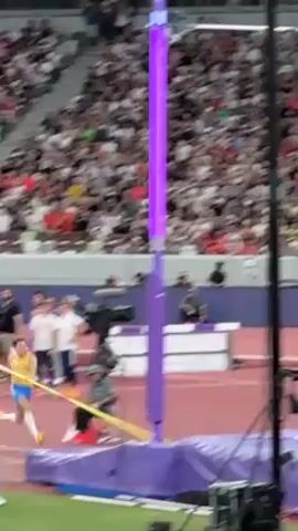 The video shows a male pole vaulter jumping during an event. He is wearing a yellow and blue uniform. The pole is yellow and the standards are purple.  He successfully clears the bar, but lands on his back on the mat. He sits up almost immediately and appears to be uninjured. The event is taking place in front of a large crowd in a stadium. A large ad reading “Sound Mind, Sound Body” is visible on the banner behind the standards, as is the URL “www.asics.com”. Several flags are visible along the stadium’s roofline.