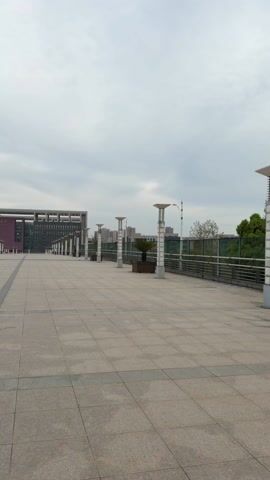 The video shows a wide, tiled walkway leading toward a large, modern building under a cloudy sky. The walkway is lined with tall, cylindrical light fixtures and planters containing small trees. Two people are walking in the distance towards the building. A thin, dark gray line runs down the center of the walkway. Other buildings and trees can be seen in the background.