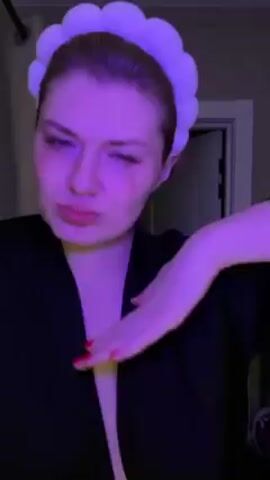 A woman with a white headband and wearing a black robe or jacket is shown in a close-up shot. The lighting is a consistent purple hue, giving the video a distinctive color cast. The woman makes various facial expressions, including a frown, a smile, and a pout. In the background, a white door is visible. The overall scene appears to be set in a bathroom or similar personal space.