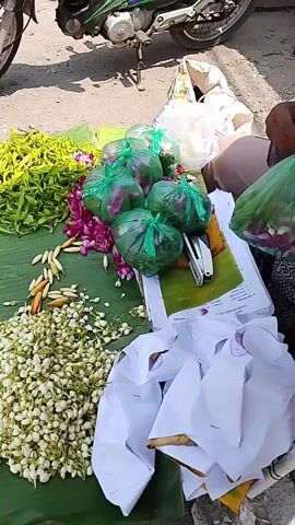 The video shows a close-up, top-down view of a market stall. The stall is covered with a large green leaf, likely a banana leaf, which serves as a base for displaying various items. In the foreground, to the left, is a large pile of small, white, unopened flower buds, possibly jasmine. To the right of the flower buds, there are some long, slender yellow seed pods or perhaps dried flowers arranged in a line. Further back, on the green leaf, there are several purple and pink flowers, some of which appear to be lotus flowers or similar. Arranged on top of these are several items wrapped in translucent green plastic bags, tied with green string. Each bag appears to contain multiple round, reddish-purple objects, possibly fruits or vegetables like onions or shallots. Behind these green bags, there is a stack of papers or books, one of which has a picture of green foliage on it. To the far right of the frame, a person's hands are visible, dressed in a patterned cloth, reaching towards one of the green plastic bags and the paper beneath it. The person appears to be interacting with the items on the stall. In the background, a motorcycle is partially visible. The ground around the stall appears to be asphalt or a similar paved surface. There are also several white paper bundles, tied with string and some with gold trim, scattered around the front and right side of the stall. The overall impression is of a flower and produce stall at an outdoor market.