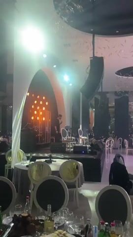 The video shows a man standing on a stage at an event, looking down and to his left, with his hand on his stomach. The stage has a white and black backdrop with an orange arch and several lights. There are chairs, microphones, and sound equipment on the stage. The hall is decorated with tables and chairs for guests. The ceiling has ornate designs and lighting fixtures. There are people at the tables. The man begins to perform as other people dance in the room.