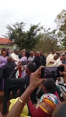Here is a description of the video:

The video captures a group of people gathered in an outdoor setting. The crowd includes men in black suits, individuals in religious robes, and women in colorful clothing. Many members of the crowd are using their phones to record or take pictures, aiming them towards the center of the group. The setting appears to be a casual gathering, possibly after a formal event or ceremony. In the background, there is a building with a red roof and some trees. The overall atmosphere seems celebratory, with music playing in the background.