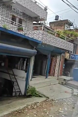 The video pans across a street in a town or city. Buildings line the street, some with multiple stories. They appear to be a mix of commercial and residential properties. Electrical wires are strung haphazardly between poles and buildings. There are several parked vehicles including cars and motorcycles. Pedestrians are visible on the sidewalks and walking in the street. Some of the buildings have shops on the ground floor with merchandise displayed outside. Trash is visible on the ground in some areas. The weather appears to be overcast or cloudy. The overall impression is of a busy, somewhat cluttered urban environment.