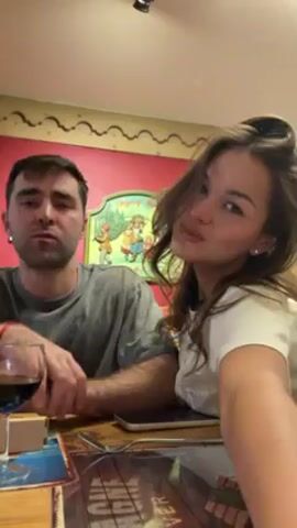 Here's a description of the video:

A young couple is sitting at a wooden table. The man is on the left side of the frame, wearing a gray shirt and holding a glass of red wine. The woman is on the right, wearing a white shirt with her hair styled.  She is recording the video. The woman leans over and kisses the man twice on the lips.  Behind them is a wall painted partly red, with decorative artwork on it.  There's a smartphone and silverware on the table in front of them.
