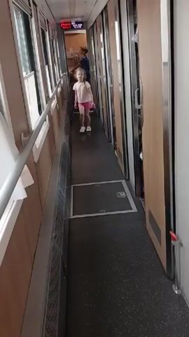 The video shows a young girl walking down a train hallway. She is wearing a pink t-shirt and pink shorts. She has blonde hair and is wearing white shoes. In the background, a woman in a blue dress is walking further down the hallway. The hallway is narrow and has doors on either side. The floor is dark gray and the walls are a light beige color. There are handrails on the left side of the hallway.