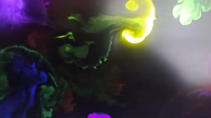 This is a close-up, abstract shot of colorful smoke or liquid inks swirling and mixing against a dark background. The colors include bright yellow, neon green, vibrant purple, and deep blues and reds. The smoke trails create organic, fluid shapes that resemble nebulae or paint splatters. The lighting highlights the luminescence of the colors, creating a glowing effect. There are no people, objects, or discernible actions other than the natural movement of the colored substances. The environment appears to be a liquid medium or space where these colorful elements can freely interact and dissipate.
