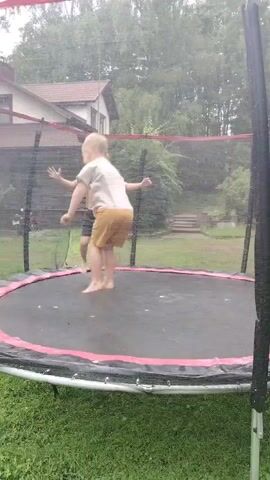 Two boys are jumping on a trampoline in a backyard. One boy is wearing a white t-shirt and yellow shorts. The other boy is wearing a yellow t-shirt and black shorts. It appears to be raining, as there are water droplets visible in the air and on the trampoline. The trampoline is surrounded by a safety net. In the background, there is a house and trees.