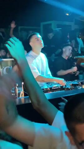 A person wearing a white t-shirt is standing behind a DJ booth, interacting with the DJ equipment. There is a person with blonde hair in a black top to the left of the DJ, and another person in a black t-shirt to the right. Several other people are visible in the background, some with their hands raised, suggesting they are at a nightclub or similar event. The lighting is dim with blue and white spotlights.