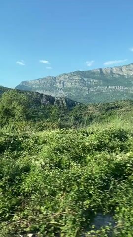 The video shows a view from a moving vehicle traveling through a mountainous, green landscape under a clear blue sky with a few white clouds. In the foreground, there is dry grass and rocky terrain. Further back, lush green vegetation covers the hillsides, and some trees, including what appear to be cypress trees, are visible. In the middle ground, several small houses with red roofs are nestled among the trees. In the background, large, rugged mountains with rocky peaks dominate the horizon. The sun is shining brightly, casting light on the scene. The video appears to be shot on a sunny day, possibly during the summer or late spring, given the vibrant green foliage. The overall impression is of a scenic rural or semi-rural environment in a hilly or mountainous region.